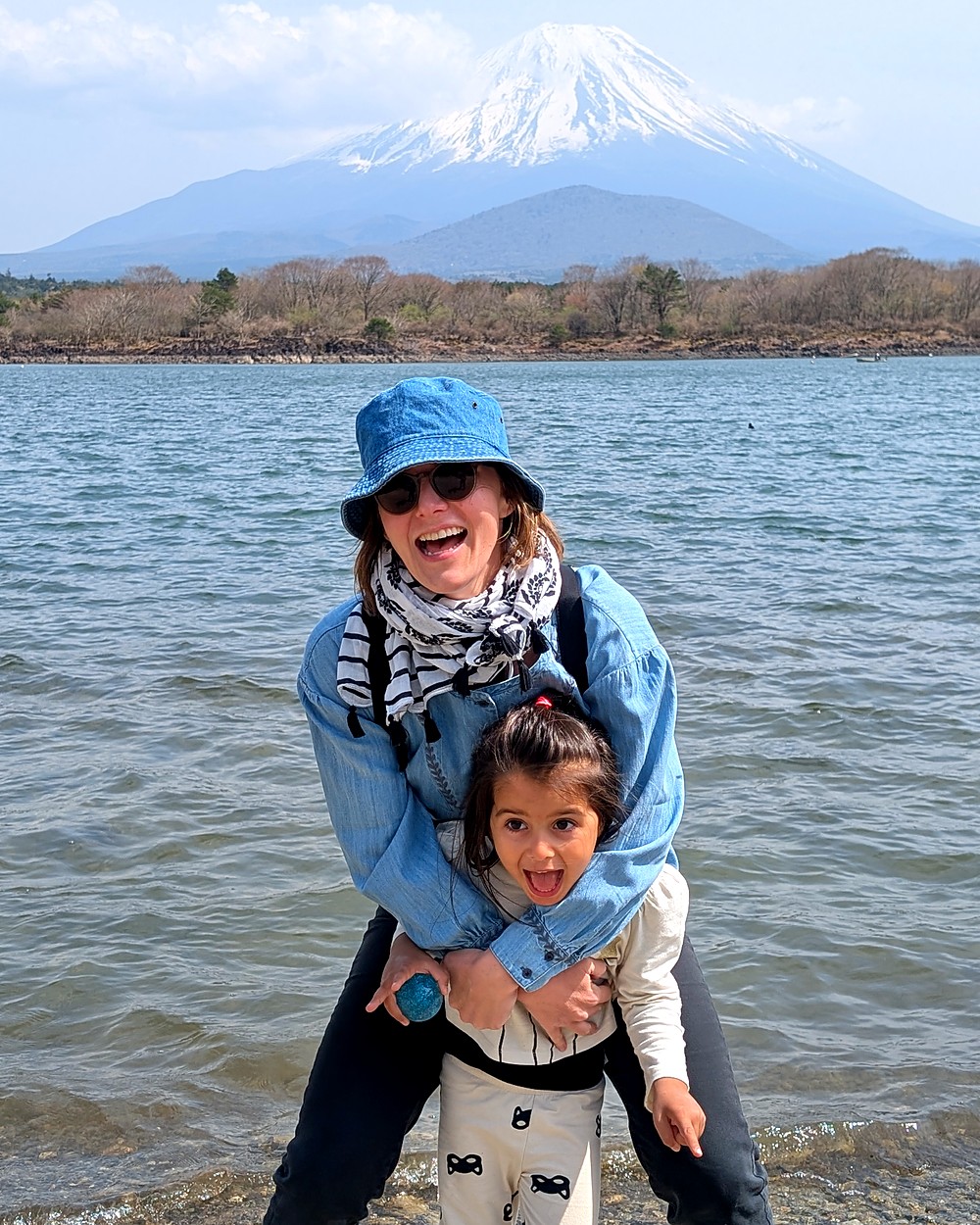 A parent and young child laughing together on the shore of Lake Shojiko, with Mount Fuji snow-capped behind them