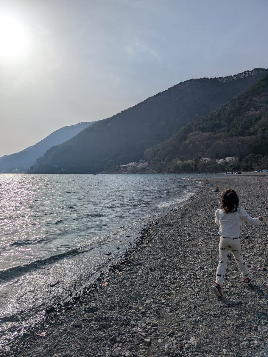 A child running along the stony shore of Lake Saiko in the late afternoon, hills rising across the water