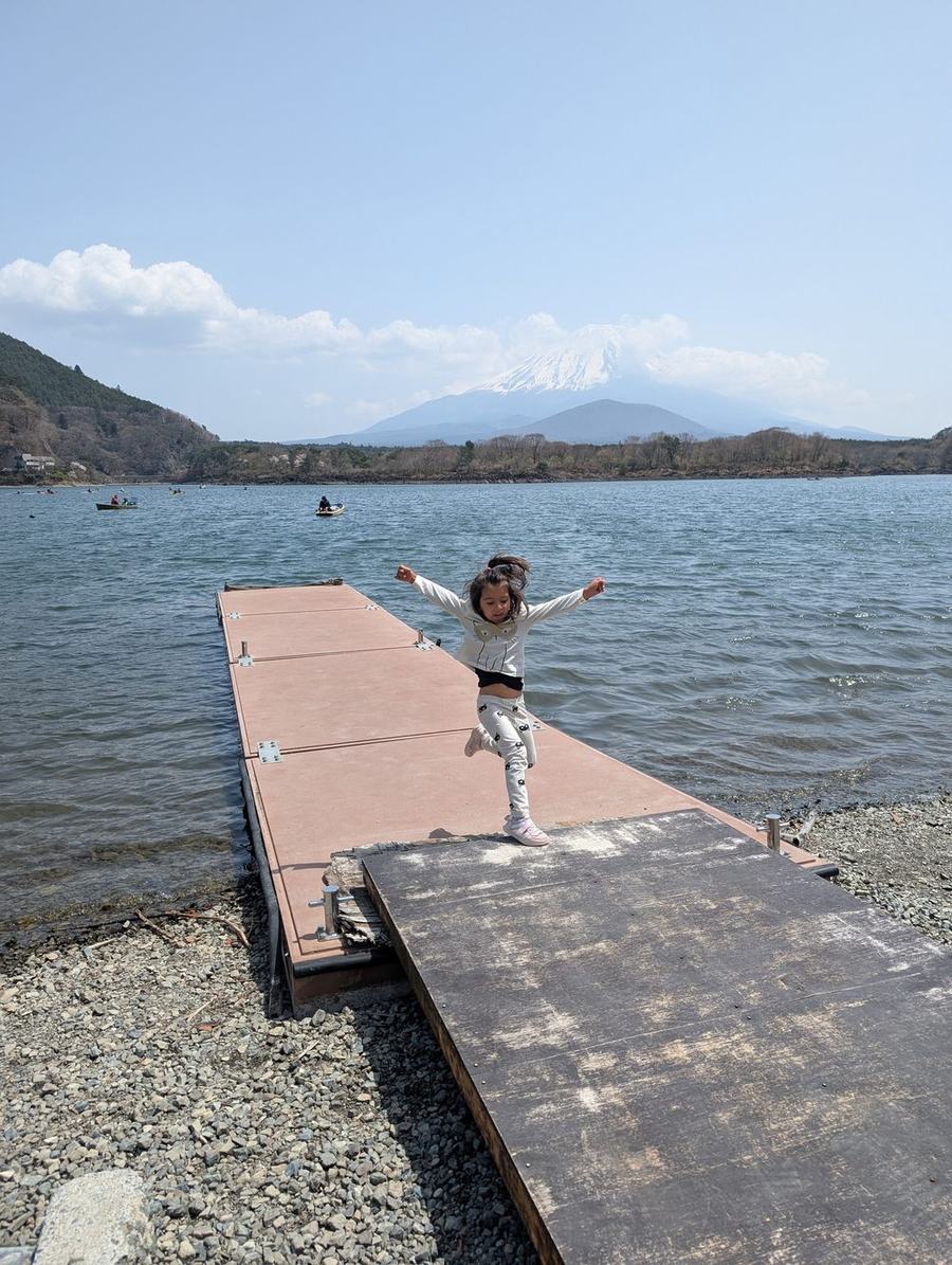 A child standing arms-out at the end of a small wooden jetty on Lake Shojiko, Mount Fuji across the water